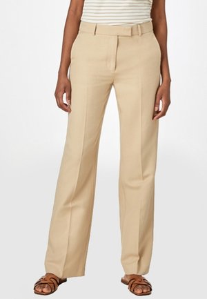 Kobieta ubrana w beżowe spodnie o prostych nogawkach, włożony do środka pasiastego topa oraz brązowe sandały z odkrytymi palcami, stojąca na tle gładkiego tła.