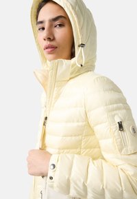 Light yellow puffer jacket with a zippered front, high collar, and detachable hood. Features ribbed texture and logo detail on sleeve.