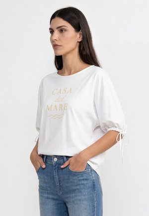 Kobieta z długimi ciemnymi włosami, w białej koszuli z napisem „CASA del MARE” i niebieskich dżinsach, dłonie w kieszeniach, patrząca w lewo, neutralne tło.