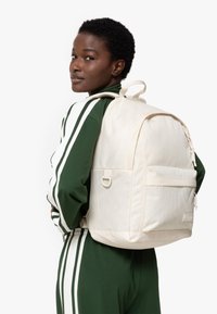 Sac à dos couleur crème en tissu texturé, présentant une poche zippée à l'avant, des détails de couture et des bretelles ajustables.