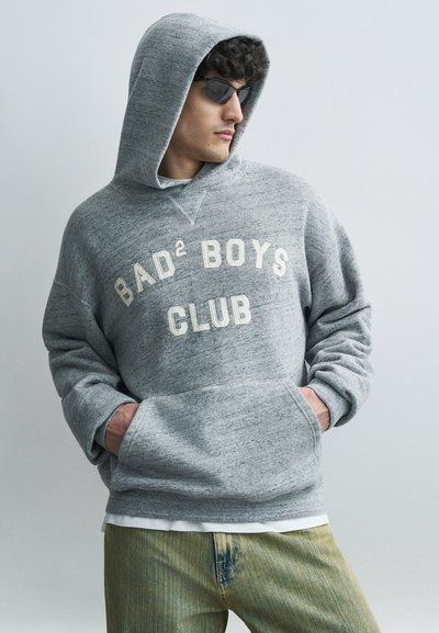Homme portant un sweat à capuche gris avec le texte "BAD² BOYS CLUB", des lunettes de soleil, et un pantalon vert jaunâtre, les mains dans les poches du sweat.