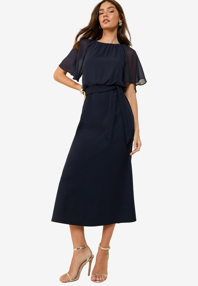 Marineblaues Kleid mit transparenten, schwebenden Ärmeln, einem taillierten Bindegürtel und einem fließenden Maxi-Längen-Schnitt. Kombiniert mit silbernen, geflochtenen High Heels.