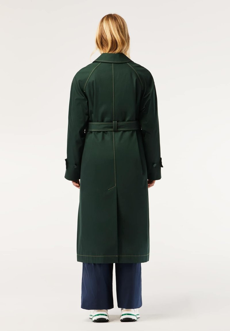 Manteau long vert avec une taille ceinturée, comportant deux poches latérales et des coutures jaunes contrastées. Tissu lisse avec un design de col classique.