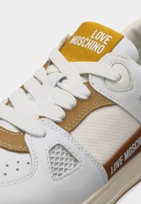 Λευκό και καφέ sneaker Love Moschino με λευκά κορδόνια και λεπτομέρεια από δίχτυ, με εμφανές το όνομα της μάρκας στη γλώσσα και στο πλαϊνό λουράκι.