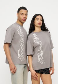 Dos modelos están uno al lado del otro vistiendo camisetas grises oversize a juego con texto en script vertical blanco que dice "Furnished Idea."