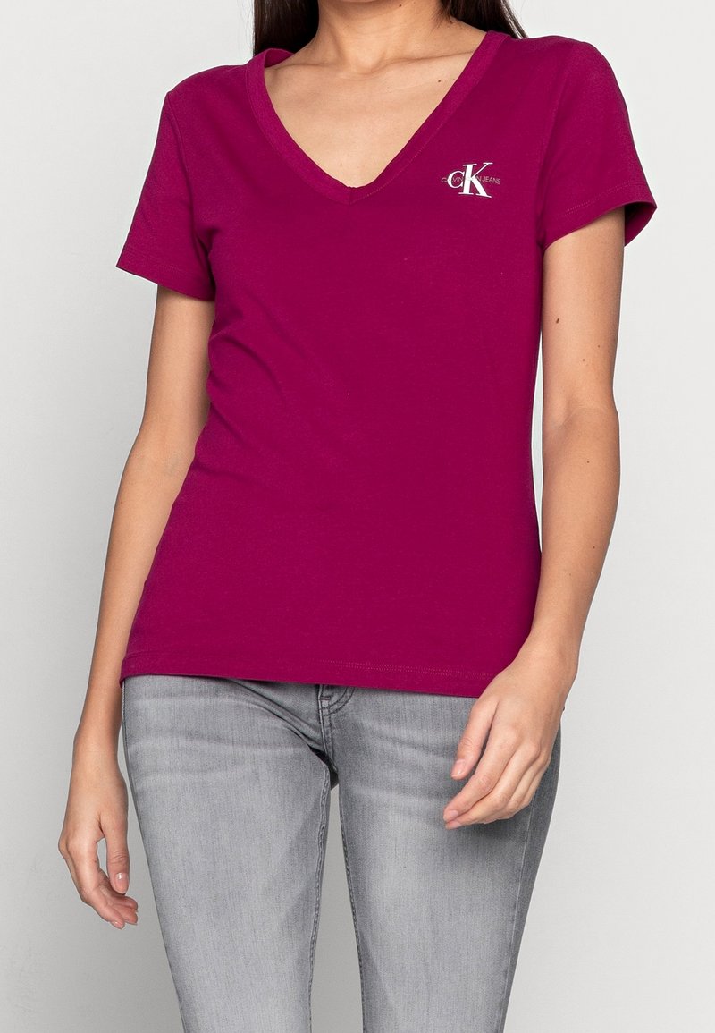 Magenta V-Ausschnitt T-Shirt aus Baumwolle, mit kurzen Ärmeln und einem kleinen Logo auf der linken Brust. Kombiniert mit grauen Jeans.