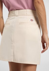Beige kjol i slätt tyg, med två bakfickor, detalj med knappar och en Dickies-logotyp på höger sida.