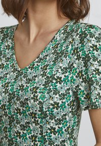 Robe fleurie avec un décolleté en V, en tissu texturé vert clair, vert foncé et fleurs jaunes sur un fond pâle. Manches courtes.