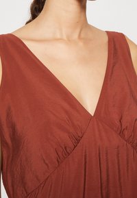 Robe couleur rouille avec un décolleté en V profond, tissu froncé au niveau de la poitrine, et matériau lisse légèrement texturé.