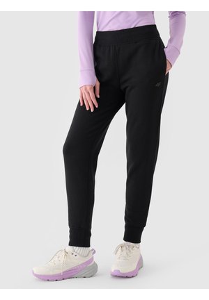 Persona que lleva pantalones deportivos negros, zapatillas blancas con detalles en púrpura y una camiseta de manga larga color lavanda, de pie frente a un fondo liso.