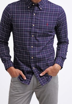 Homme portant une chemise à carreaux bleu foncé boutonnée avec des lignes violettes et blanches, les mains dans les poches, associée à un pantalon gris clair.
