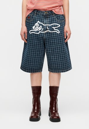 Άτομο που φοράει μπλε τζιν σορτς με σχέδιο λευκής καρικατούρας γάτας, κόκκινη και λευκή ριγέ μπλούζα και σκούρα κόκκινα μπότες μέχρι τη μέση της γάμπας.