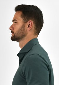 Donkergroene polo met een kraag, met een gladde textuur en een getailleerd ontwerp, getoond op een mannelijk model in profiel.