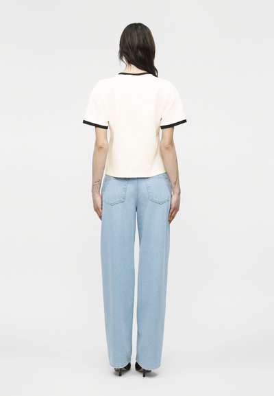 Femeie cu păr lung și întunecat, văzută din spate, purtând un tricou alb cu mâneci scurte și detalii negre și blugi largi, de culoare albastru deschis, cu tocuri.