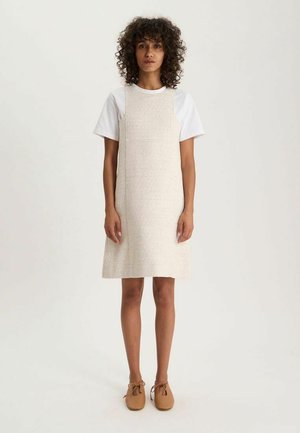 Femme aux cheveux bouclés portant une robe beige texturée sans manches sur un t-shirt blanc et des chaussures à lacets marron clair, debout devant un fond uni.