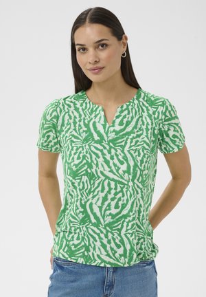 Kvinde med langt, mørkt hår iført grøn og hvid mønstret kortærmet top og blå jeans, stående med hænderne bag ryggen mod en hvid baggrund.