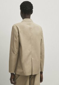 Veste en mélange de coton beige, design structuré, col à revers, fente au dos pour un meilleur mouvement, manches longues, texture lisse, style minimaliste.