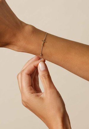 Zarter Goldkettenarmband mit horizontalem Kreuzanhänger am Handgelenk, das von einer Hand vor einem einfachen beigen Hintergrund befestigt wird.