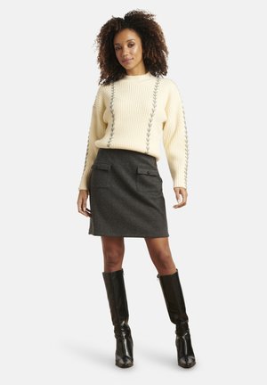 Crèmekleurige gebreide trui met geribbelde structuur en zijdetails, gecombineerd met een donkergrijze rok met twee voorzakken en zwarte kniehoogte laarzen.