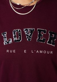 Bordeaux shirt met het woord "LOVER" in zwarte pailletten, met daaronder in witte letters "RUE E L'AMOUR" gedrukt. Textuurstof, casual ontwerp.