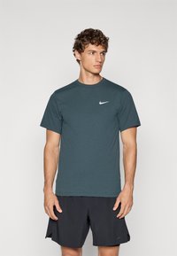 Mørk teal Nike kortermet treningstrøye i glatt stoff, med en hvit logo på brystet, sammen med svarte shorts.