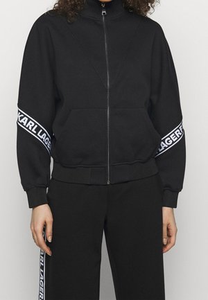 Czarna kurtka zapinana na zamek z białymi paskami z napisem "KARL LAGERFELD" na rękawach, zestawiona z dopasowanymi czarnymi spodniami z bocznymi paskami z logo.