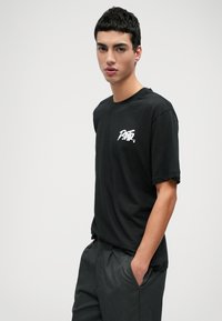 Μαύρο κοντομάνικο μπλουζάκι με λευκό γραφικό λογότυπο στο αριστερό στήθος, σε συνδυασμό με σκούρες γκρι παντελόνες. Λείο ύφασμα, άνετη εφαρμογή.