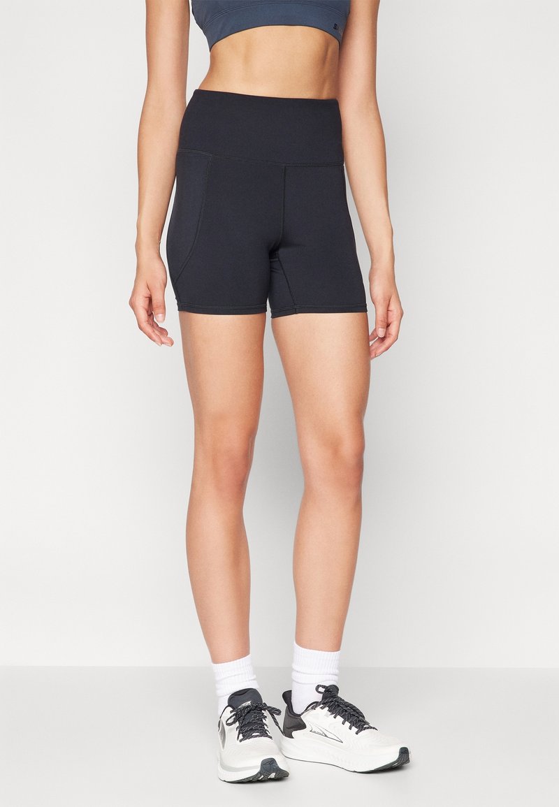 Shorts de sport noirs avec une taille haute, dotés de deux poches latérales et de coutures contrastantes. Portés avec un soutien-gorge de sport gris et des baskets.