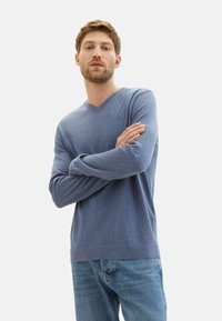 Maglione azzurro lavorato a maglia con scollatura a V, maniche lunghe e polsini e orlo a coste, abbinato a jeans azzurri chiari. Texture morbida, design semplice.