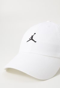 Căciulă albă din țesătură texturată, având un logo Jumpman brodat în negru pe partea din față. Brăcătură curbată cu detalii cusute.