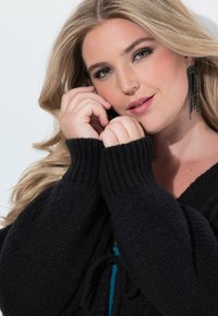 Svart stickad tröja med ribbade ärmslut och en knyt detalj; modell som bär långa, glittriga svarta örhängen, lätt smink och vågigt blont hår.