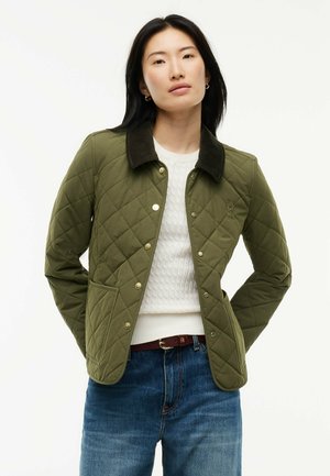 Mujer con chaqueta acolchada verde oliva con cuello oscuro, suéter blanco texturizado, vaqueros azules y pequeños pendientes de aro, de pie con las manos detrás de la espalda.