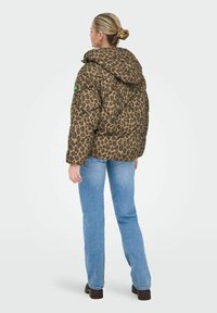 Leopard print puffer jacket with a hood, featuring a quilted design. Paired with light blue jeans and black footwear. Green logo detail.