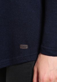 Marineblauwe gebreide trui met geribde manchetten, voorzien van een klein, bruin leren label aan de onderzoom. De stof heeft een zachte, gestructureerde uitstraling.