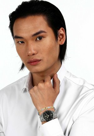 Silberne Armbanduhr mit schwarzem Ziffernblatt, Edelstahlarmband und goldenen Akzenten, getragen mit einem mehrfarbig gemusterten Armband auf einem weißen Hemd.
