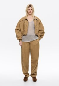 Una donna sta in piedi con sicurezza, indossando un blazer oversize beige, un top grigio lavorato a maglia e pantaloni larghi beige con scarpe marroni, su uno sfondo neutro.