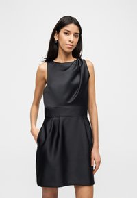 Γυναίκα που στέκεται με το ένα χέρι στην τσέπη, φορώντας αμάνικο μαύρο σατινέ φόρεμα με πλισέ φούστα και ντεκολτέ με draping, μπροστά σε μονόχρωμο φόντο.