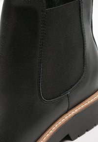 Stivale Chelsea in pelle nera con pannello elastico laterale e suola cucita marrone, in primo piano che mostra dettagli di texture e cuciture.