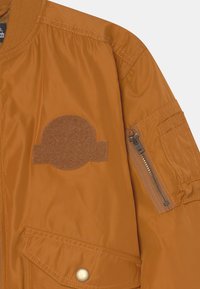 Casaco bomber em nylon laranja com um patch circular texturizado no peito, bolso com fecho no braço e bolso frontal com botão de pressão.