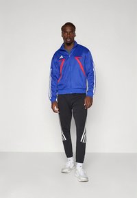 Zils rāvējslēdzēja džemperis ar sarkaniem akcentiem un baltām svītrām, kopā ar melnām biksēm, kurām sānos ir baltas svītras, un baltām sporta kurpēm.