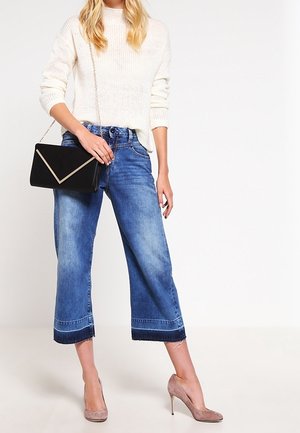 Kobieta ubrana w biały sweter, krótkie szerokie niebieskie jeansy, beżowe wysokie obcasy, trzymająca czarną kopertówkę na łańcuszku.