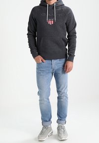 Hombre vestido con un hoodie oscuro de GANT con logo, jeans azul claros remangados en los puños y zapatillas gris claro, de pie con una mano en el bolsillo.