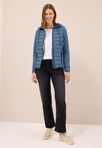 Gewatteerde blauwe jas met een capuchon, voorzien van een ritssluiting aan de voorkant en twee zijzakken, gecombineerd met een wit T-shirt en zwarte jeans.