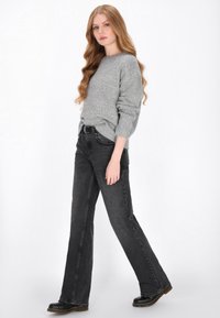Szary sweter z dzianiny z długimi rękawami, zestawiony z czarnymi dżinsami w stylu dzwonów i czarnymi butami. Modelka pozuje na jednolitym białym tle.