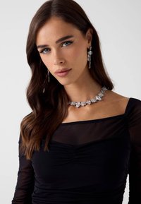 Srebrny naszyjnikkryształowy o różnych kształtach i rozmiarach, w połączeniu z pasującymi kolczykami w kształcie kropli, noszony z czarną bluzką z długim rękawem.