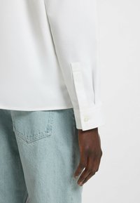 Hvid langærmet skjorte med knappet manchet, parret med lyseblå denimjeans. Skjorten har en glat tekstur og et skræddersyet design.