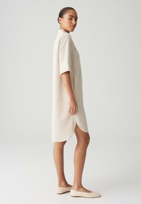 Beiges kurzärmeliges Kleid mit einem Kragen und seitlichen Schlitzen, aus einem strukturierten Stoff gefertigt. Kombiniert mit leichten, flachen Schuhen.
