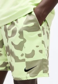Άτομο που φοράει χακί-πράσινα και μπεζ καμουφλάζ σορτς της Nike με πλαϊνή τσέπη και μαύρο λογότυπο Nike κοντά στο τελείωμα.
