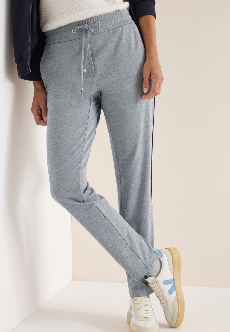 Lichtgrijze joggingsbroek met een koord in de taille, zijzakken, slim fit en donkere zijstreepjes, gecombineerd met witte sneakers met blauwe accenten.