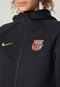 Gros plan d'une personne portant une veste noire zippée avec un logo Nike doré et un écusson coloré du FC Barcelone sur la poitrine.